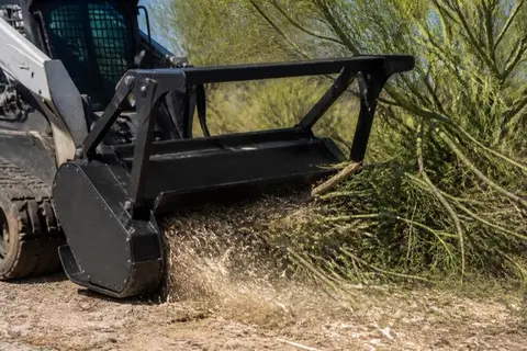 Skid steer with a mulcher head grinding volunteer mesquite on a vacant lot, chips arcing in the sunlight beside a preserved palo verde