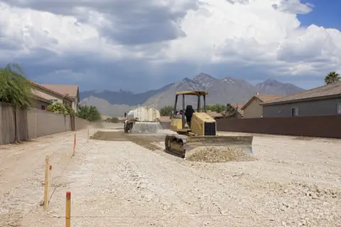 Small dozer leveling a cleared building pad with grade stakes set, water truck laying a dust-control pass behind it