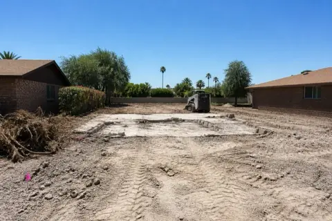 Mid-century infill lot cleared to bare pale soil with surveyor stakes and pink flagging, intact brick ranch homes on either side